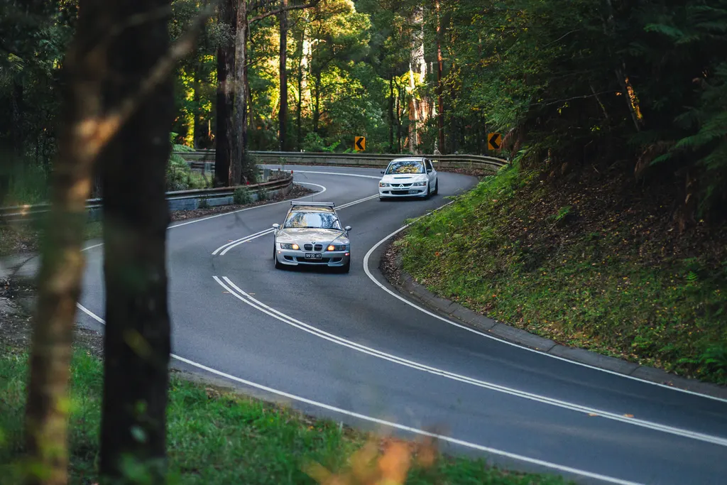 Two BMW vehicles navigating a winding forest road with dense tree coverage on both sides.