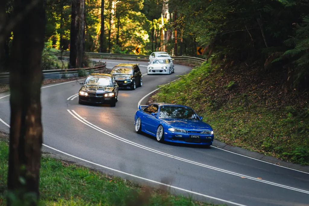 A blue sports car leads a convoy of vehicles through a forested road with dense green vegetation on both sides.