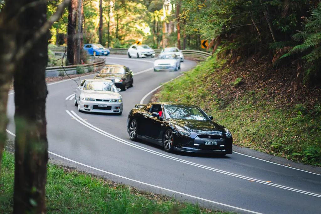 A black sports car leads a convoy of four vehicles through a tree-lined mountain road with a sharp curve.