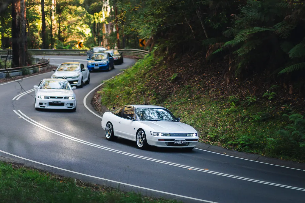A white sports car leads a convoy of vehicles through a tree-lined mountain road with sweeping curves.