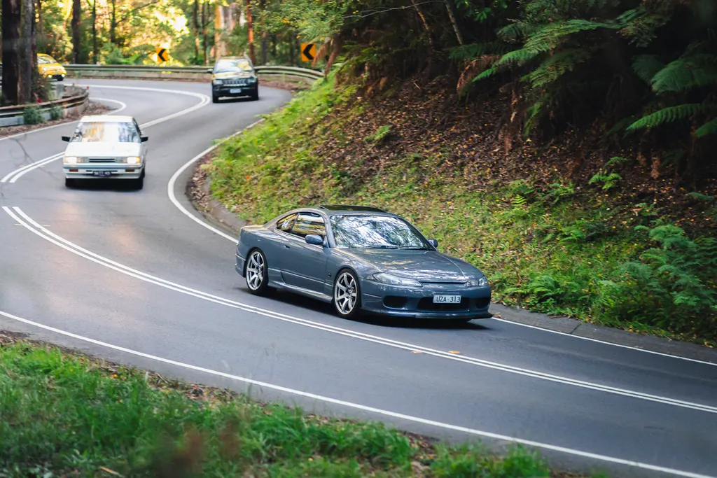Three sports cars navigating a winding mountain road through dense forest with yellow warning signs visible ahead.