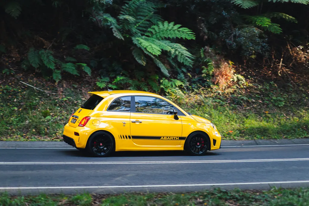 Yellow Abarth compact car parked on a road with dense green foliage in the background.
