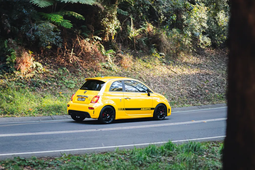 Yellow Abarth 595 compact performance car on a paved road with dense green forest in the background.