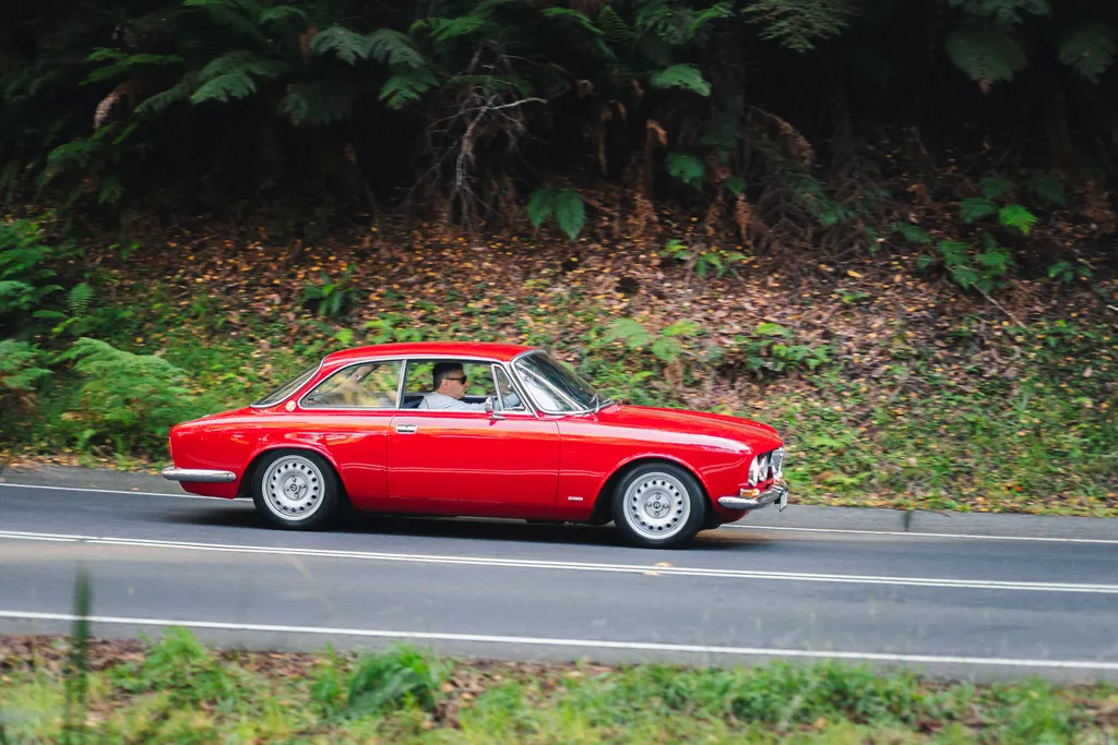 Red vintage sedan driving on a paved road with dense green forest and foliage in the background.