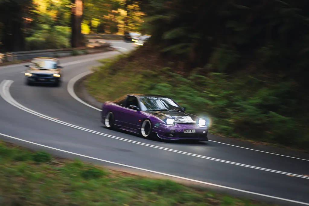 Purple sports car leading a convoy through a tree-lined road with motion blur, followed by another vehicle in the distance.