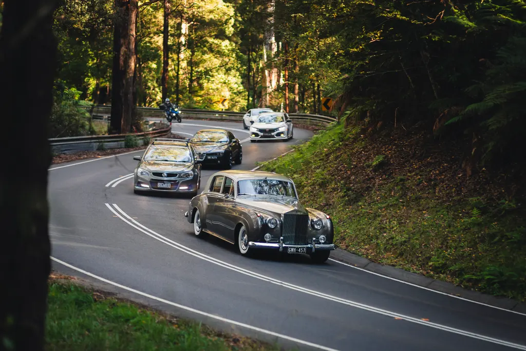 A classic black sedan leads a procession of modern cars down a tree-lined road through a forest.