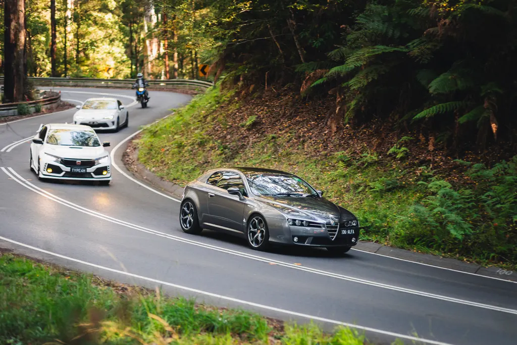 Three cars navigate a curved mountain road through dense forest canopy.