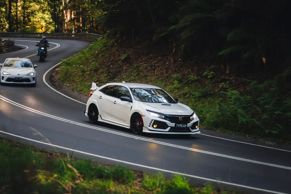 White hatchback with red accents navigates a curved mountain road lined with dense green forest, followed by other vehicles.