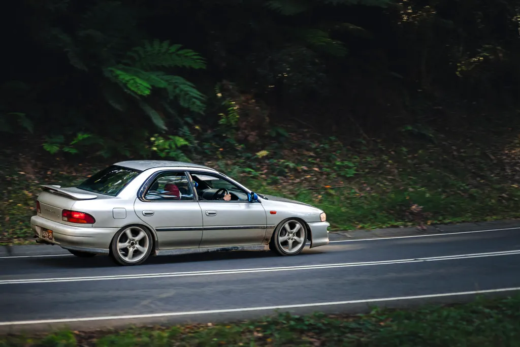 Silver sedan driving on a forested road with lush green vegetation in the background.