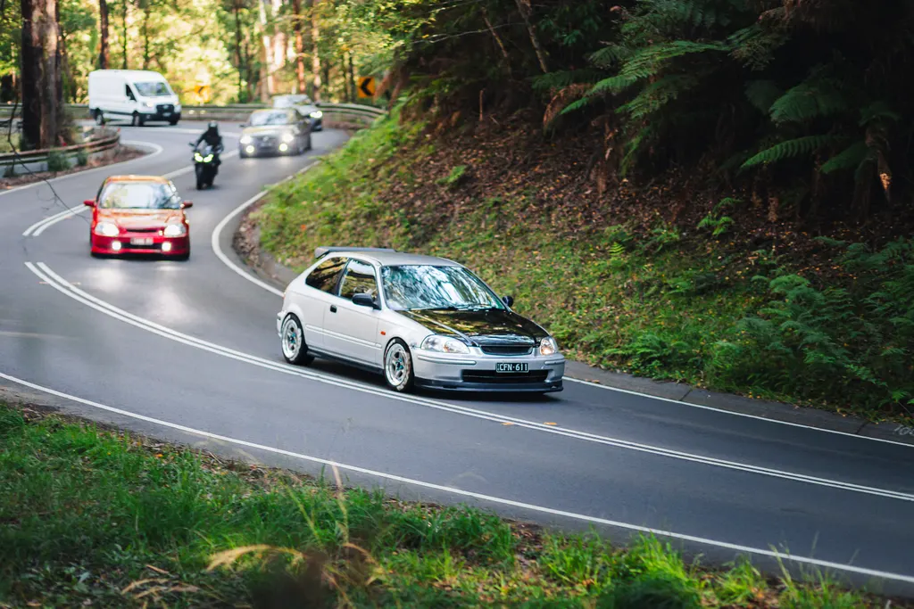 A white hatchback leads a convoy of vehicles through a forested mountain road with lush green vegetation lining both sides.