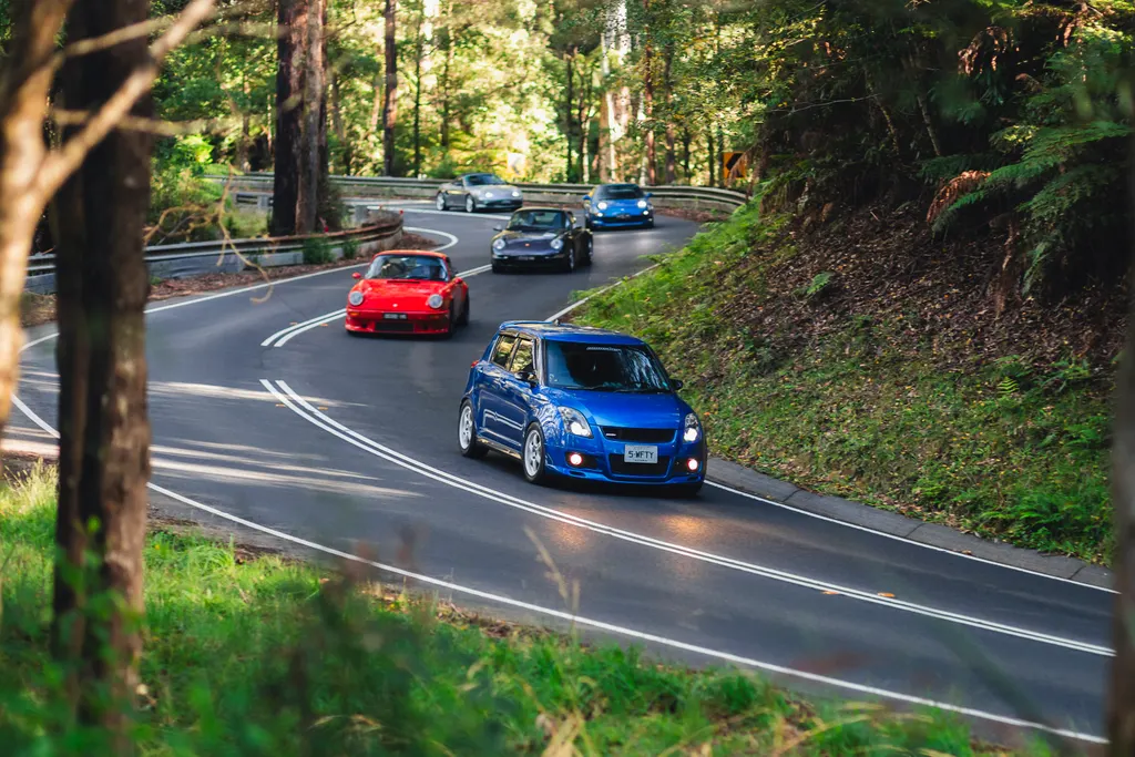 A group of sports cars navigates a winding forest road lined with tall trees and dense vegetation.