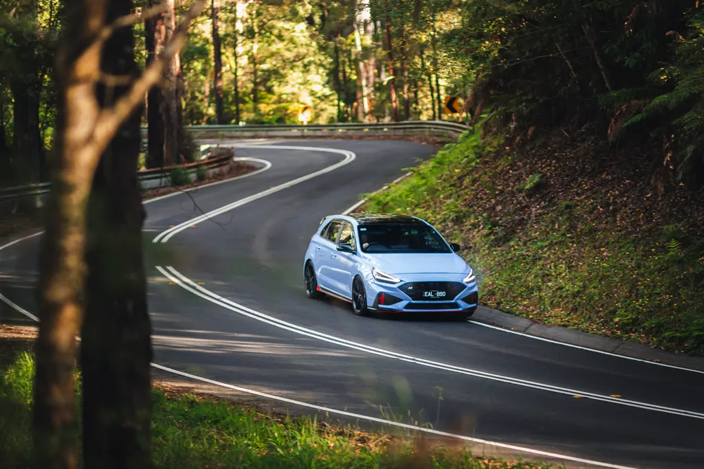 White compact car navigating a winding forest road with dense green vegetation on both sides.