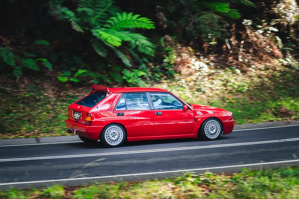 Red sedan driving on a forest road with green foliage on both sides.
