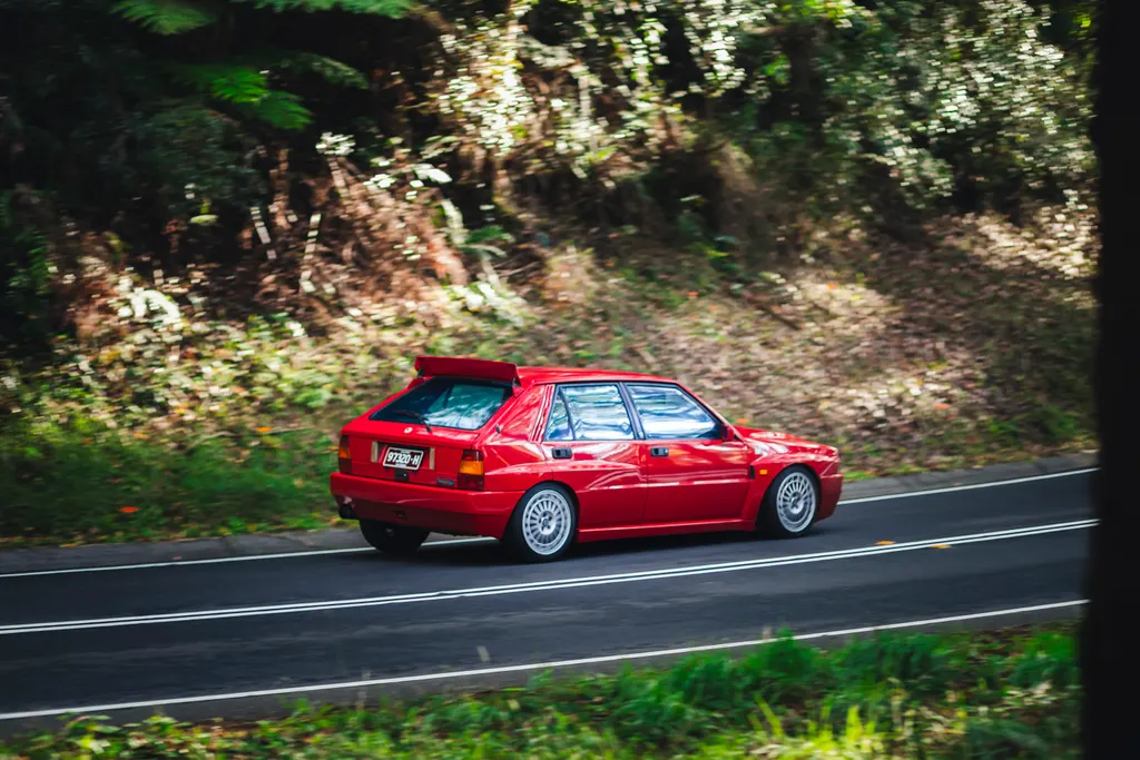 Red sedan driving on a forest road lined with green foliage and white flowering trees.