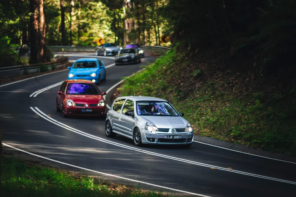 A silver Volkswagen Golf leads a convoy of compact cars through a forested road with lush green vegetation on both sides.