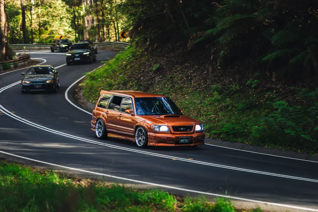 An orange SUV leads a convoy of cars through a forested mountain road with tight curves and dense green vegetation on both sides.