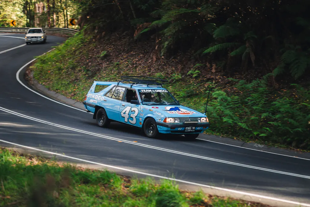 Blue race car number 43 navigating a winding forest road with lush green vegetation on the hillside.