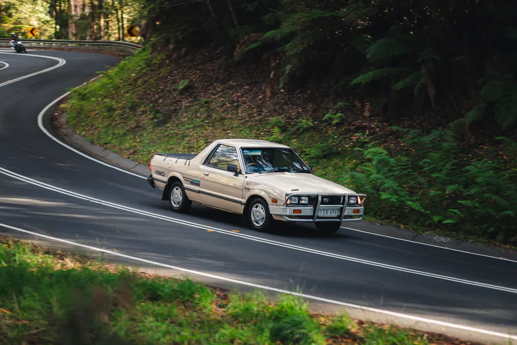 Cream-coloured sedan navigating a curved forest road bordered by dense green vegetation and moss-covered banks.