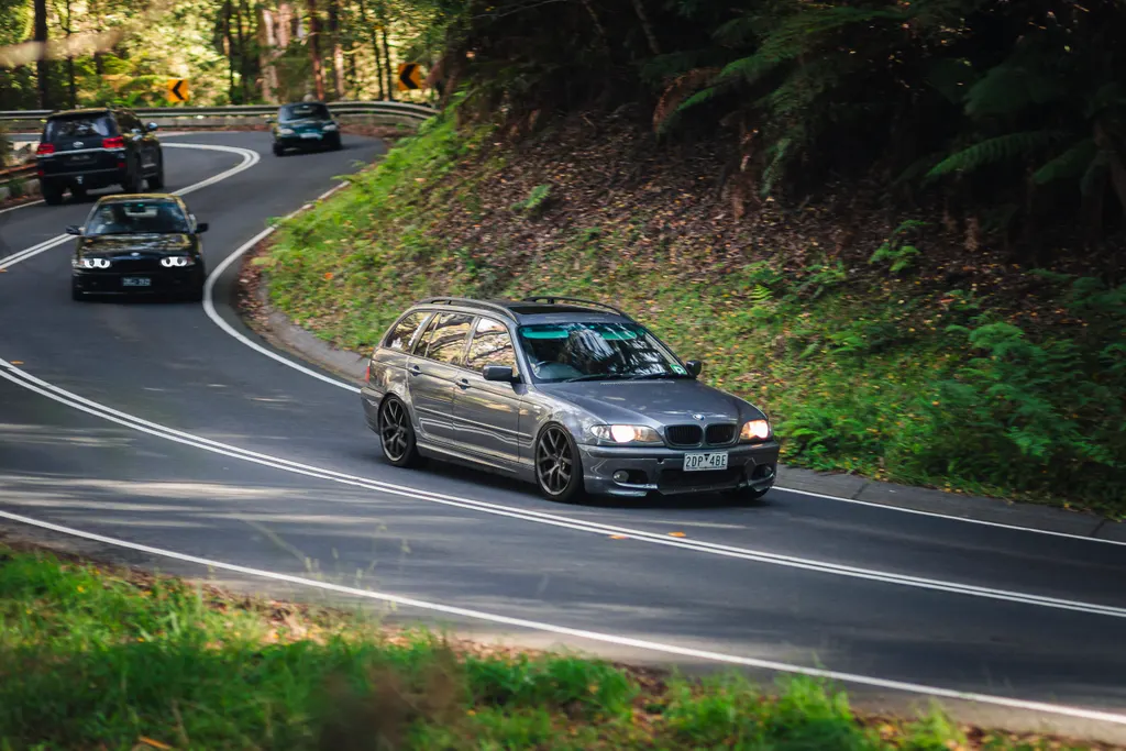 Silver sedan leading a group of cars through a wooded mountain road with tight curves and lush green vegetation.
