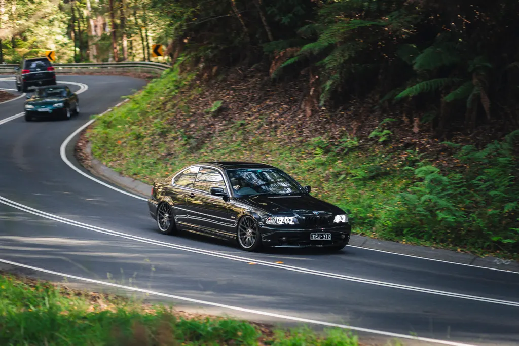Black sedan navigating a tight curve on a forested mountain road with lush green vegetation on the hillside.