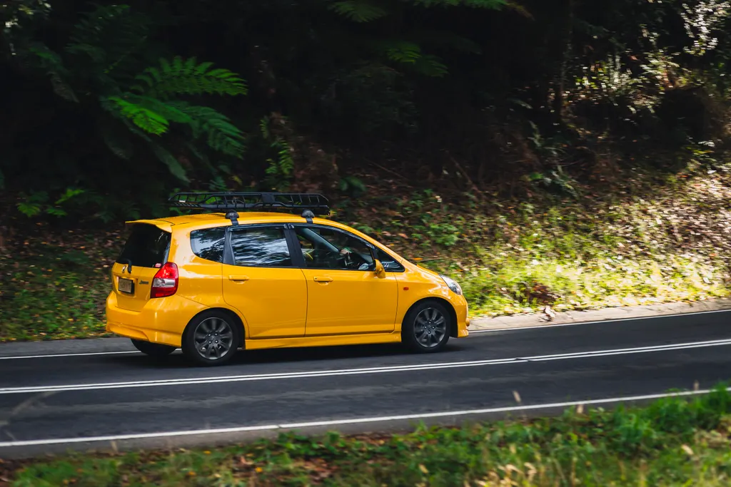 Yellow compact car with roof rack driving on a curved road lined with dense green foliage.