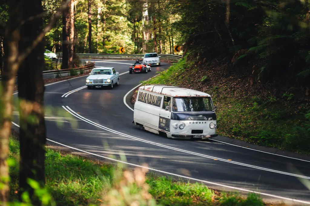 A vintage Citroën van leads a procession of classic cars through a winding forest road lined with tall trees.