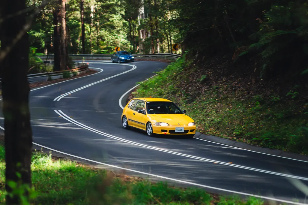 Yellow sports car navigating a curved forest road with a blue car visible in the distance.