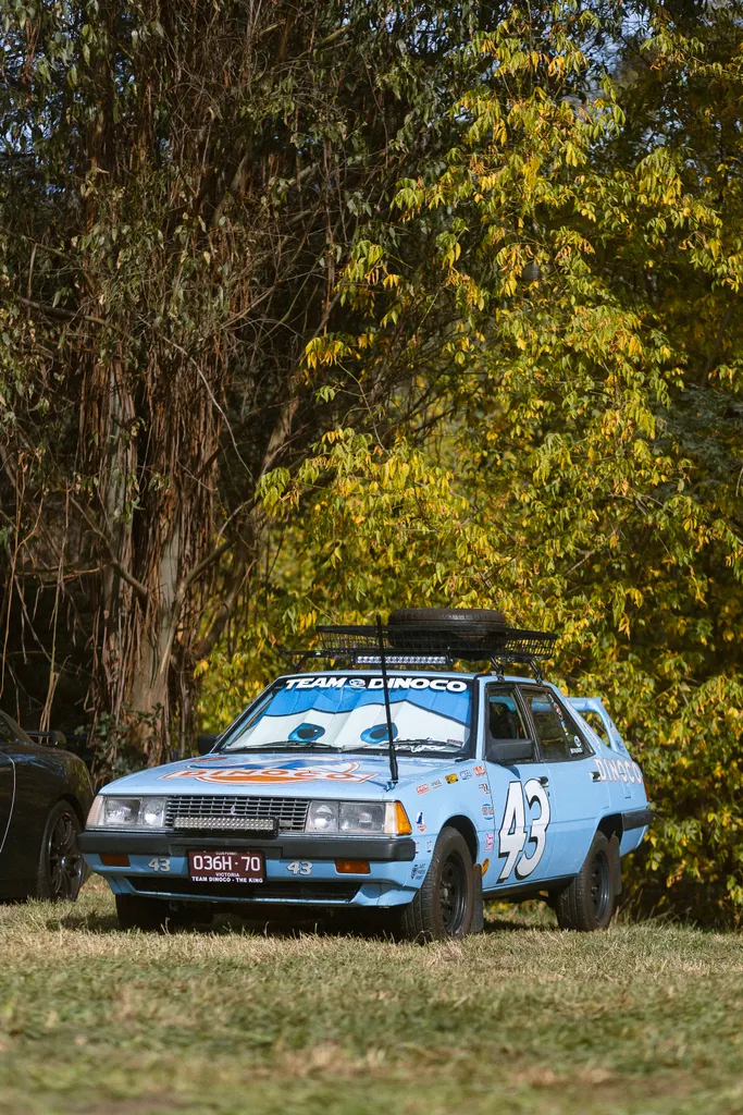 A light blue rally car numbered 43 with Team Dinoco livery parked beneath tall eucalyptus trees with autumn foliage.