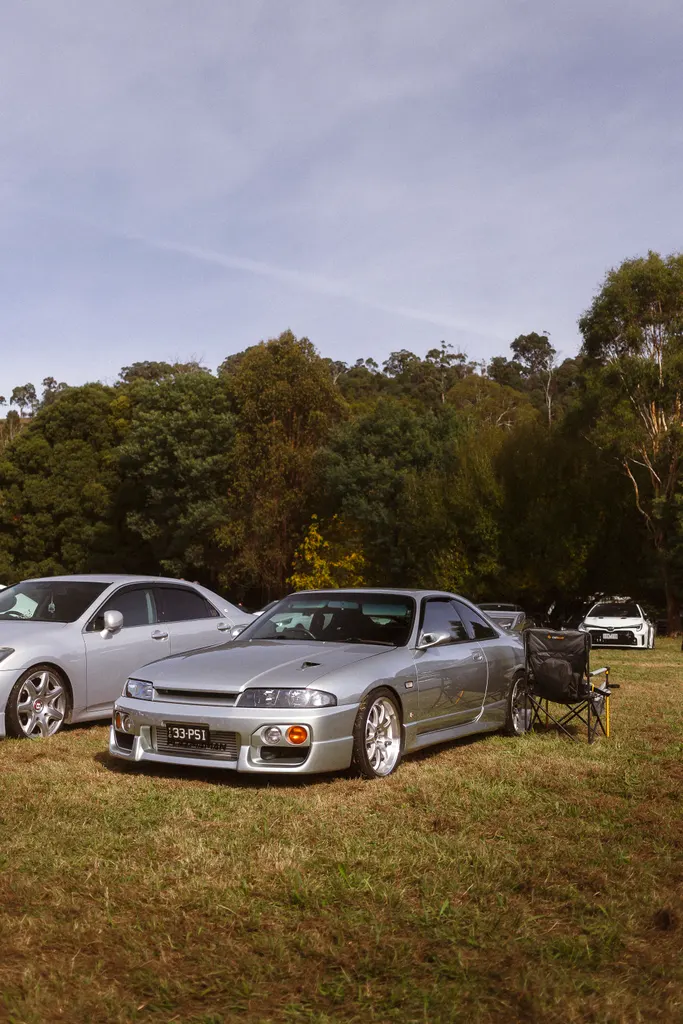 Modified silver sports car displayed at an outdoor automotive event with other vehicles and trees in the background.