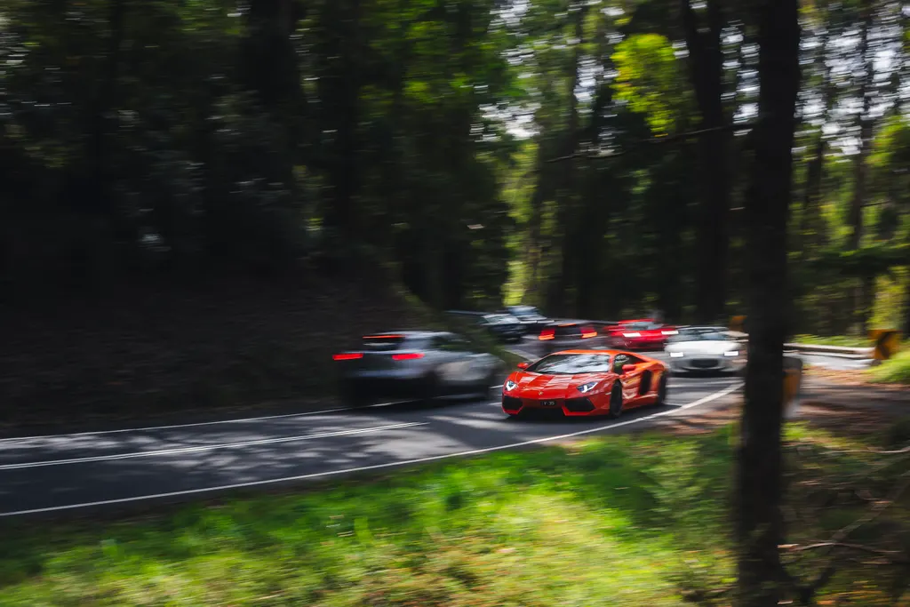 Red sports car leads a convoy of vehicles on a tree-lined road with motion blur.