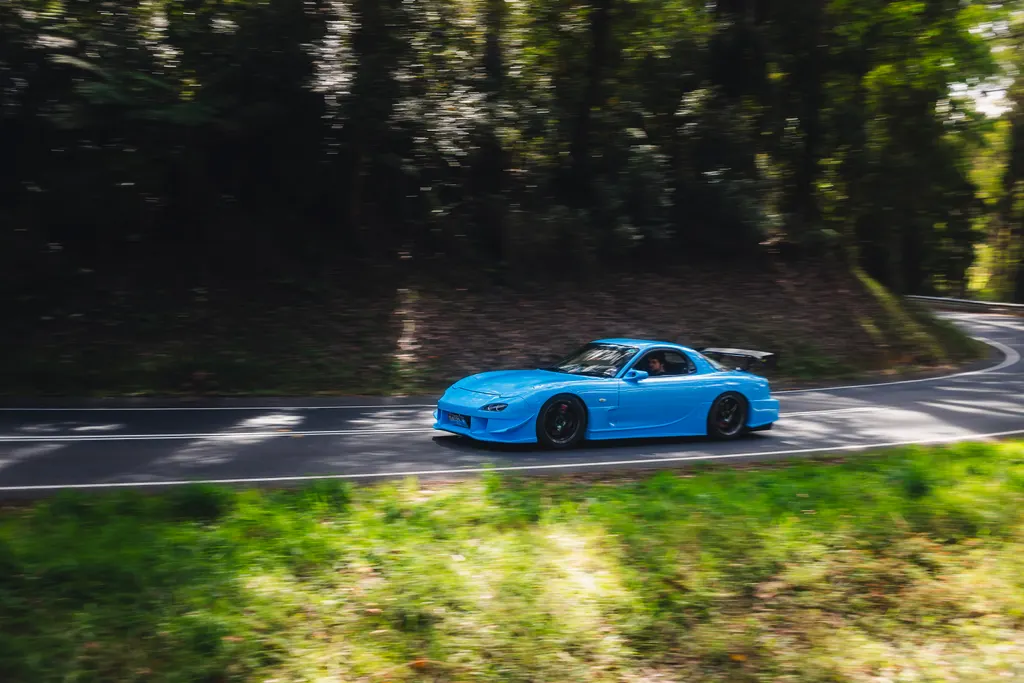 Bright blue sports car navigating a curved forest road with trees lining both sides.