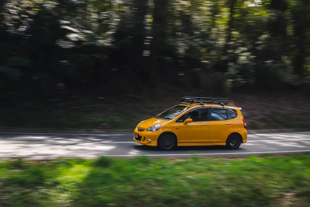 Yellow compact car with roof rack driving on a tree-lined road with motion blur from camera panning.