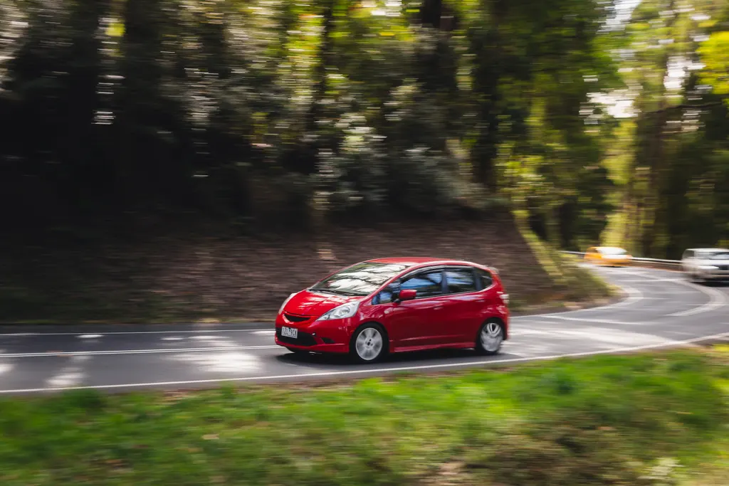 Red compact hatchback driving through a tree-lined road with motion blur in the background.