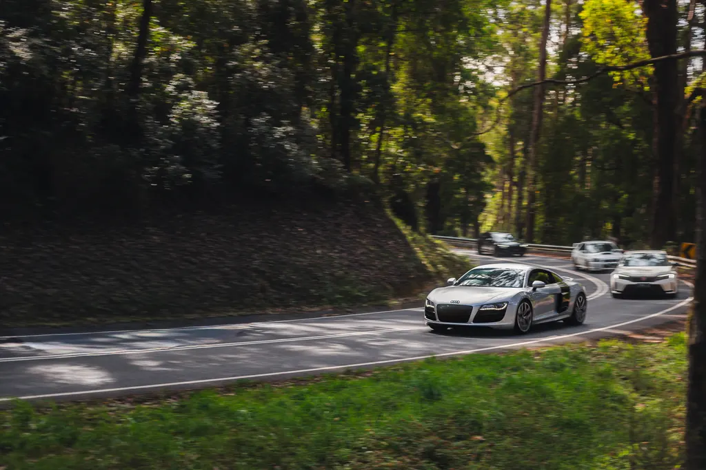 A silver Audi supercar leads a convoy of vehicles through a forested road with dense green trees lining both sides.