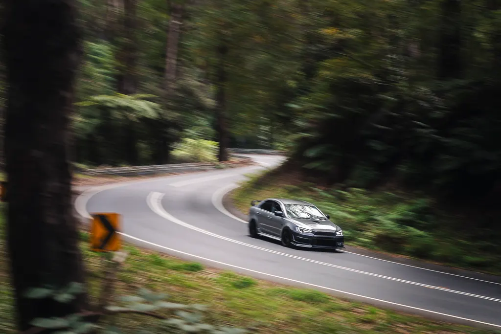 Silver sports car driving on a winding forest road with trees lining both sides.