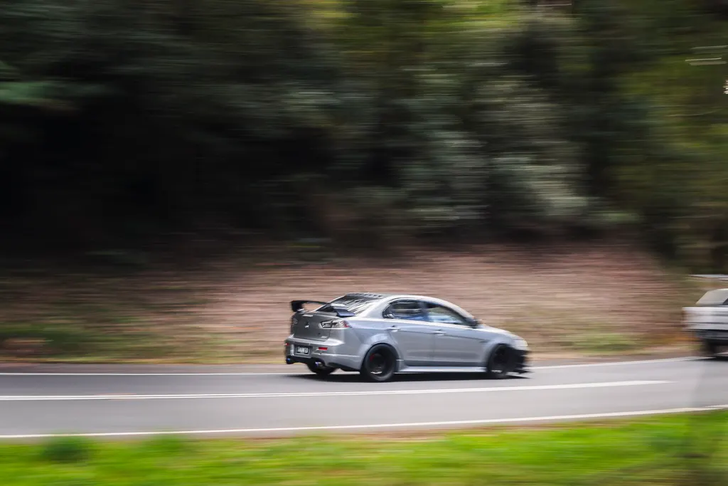 Silver compact sports sedan with rear spoiler driving on a road with blurred motion effect and green trees in background.