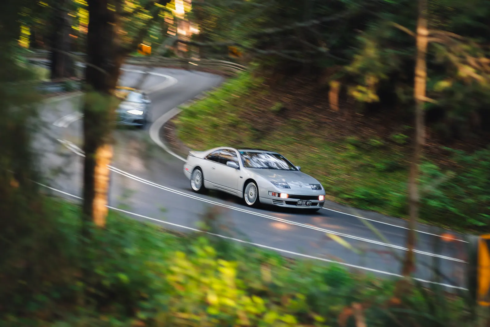 Silver sports car navigating a winding road through a forested area with trees lining both sides.