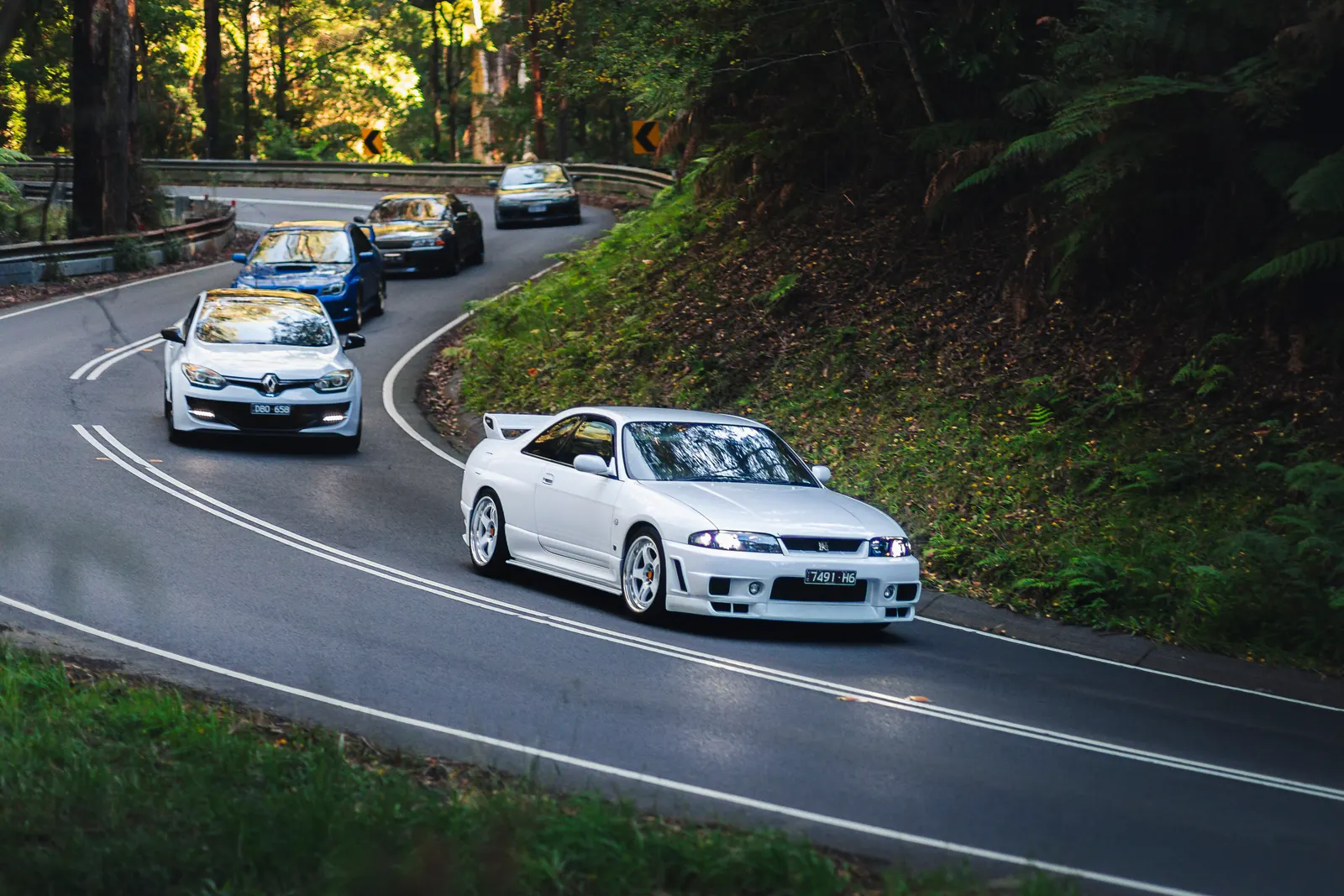 A white performance coupe leads a convoy of cars through a forested mountain road with sharp curves and dense green vegetation.