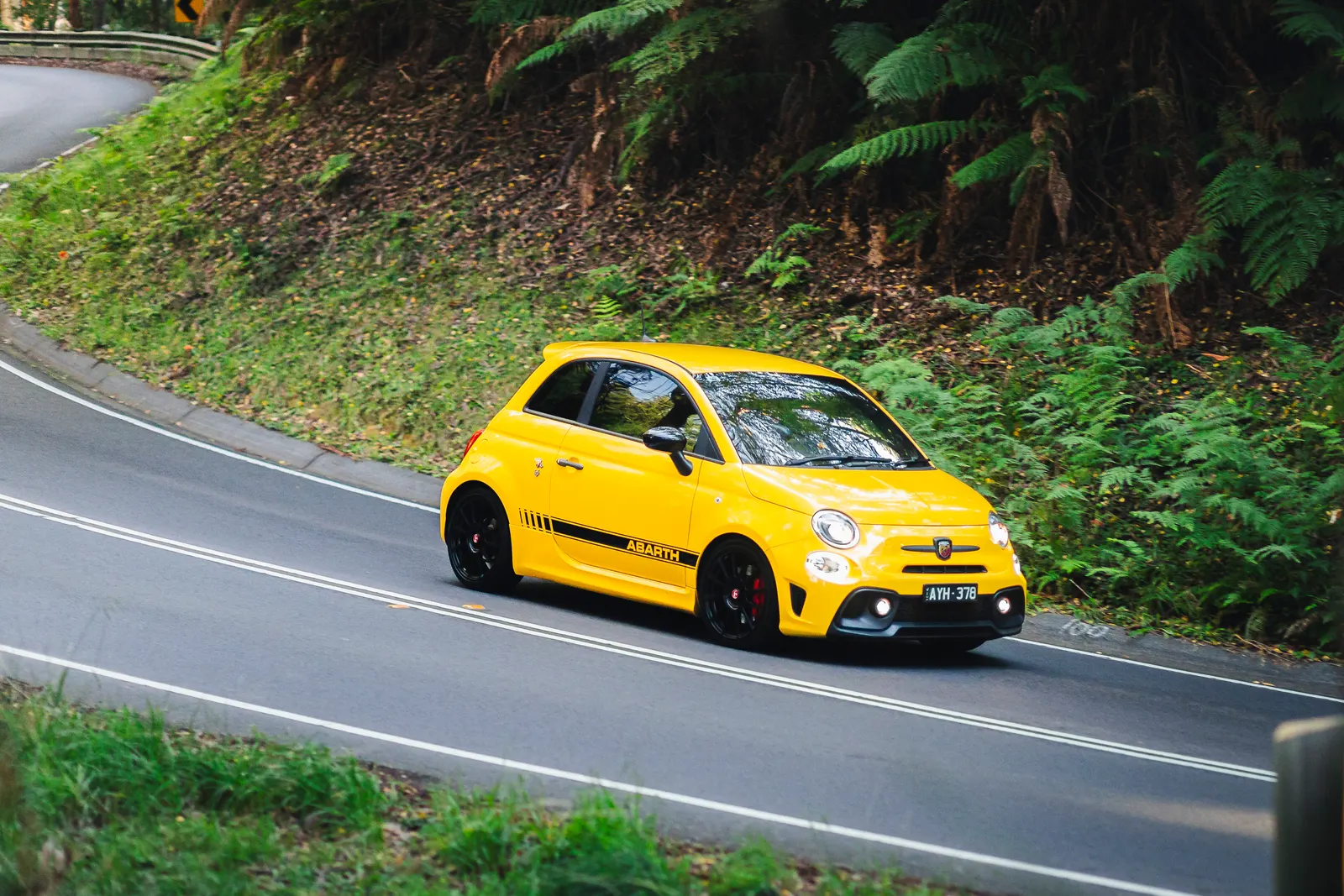 Yellow Abarth compact car navigating a winding forest road with dense green vegetation on the hillside.