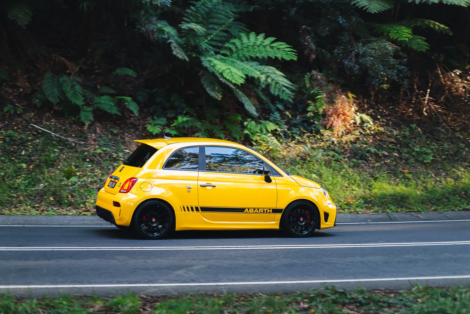 Yellow Abarth compact car parked on a road with dense green foliage in the background.