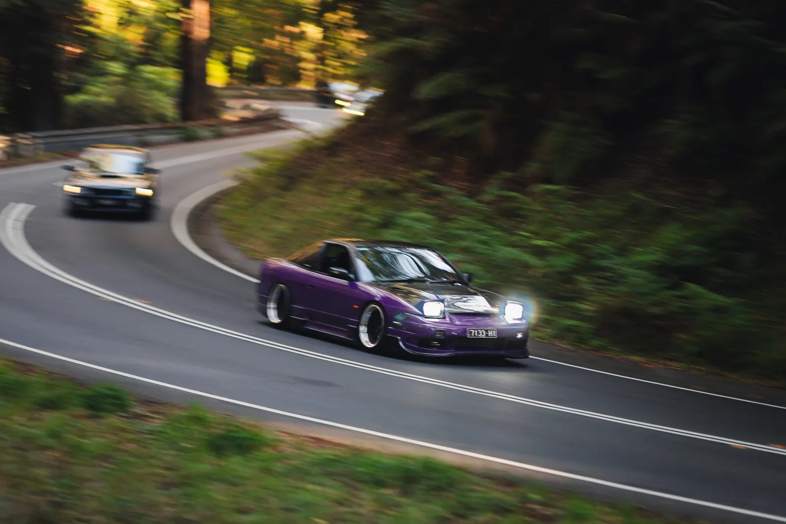 Purple sports car leading a convoy through a tree-lined road with motion blur, followed by another vehicle in the distance.