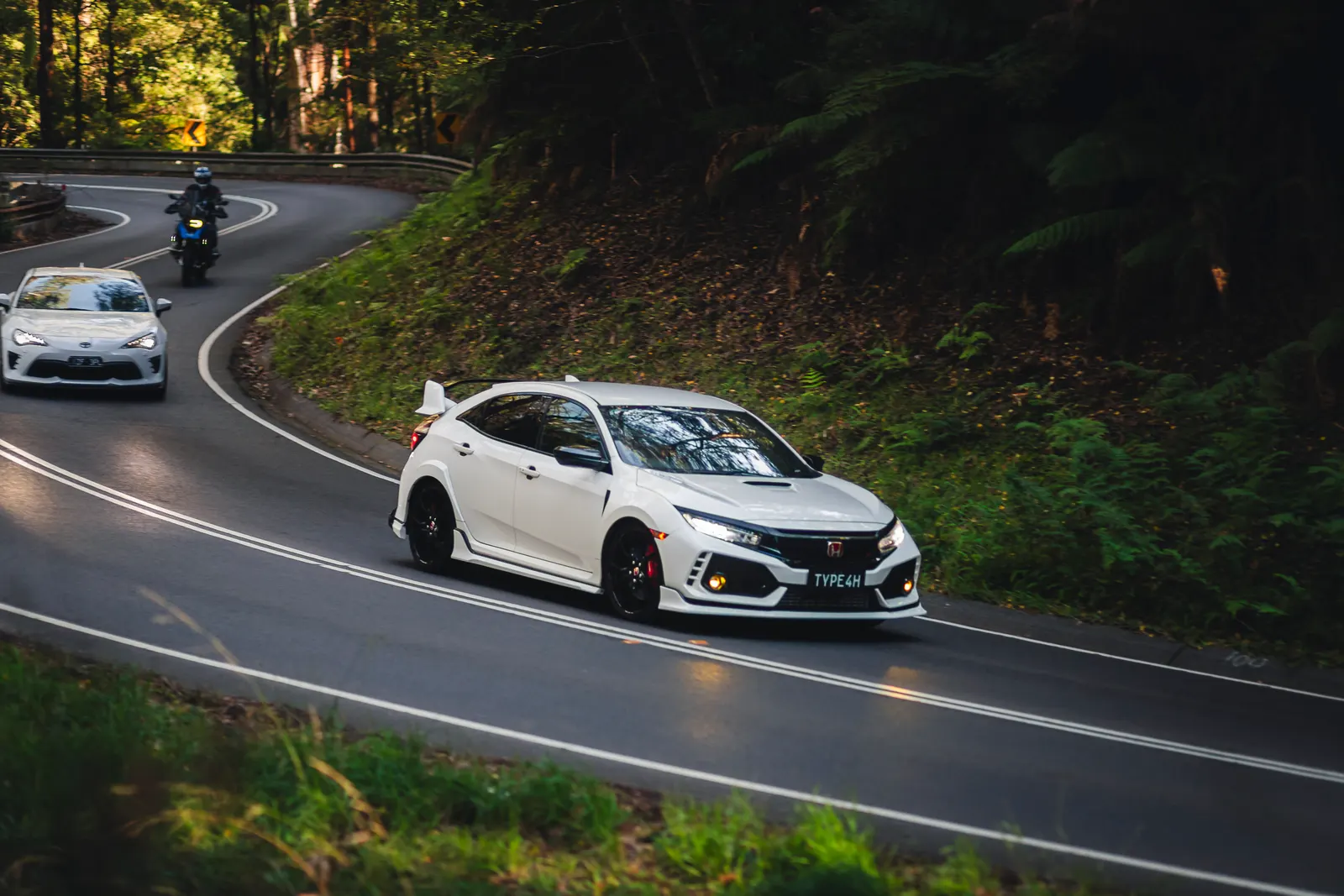 White hatchback with red accents navigates a curved mountain road lined with dense green forest, followed by other vehicles.