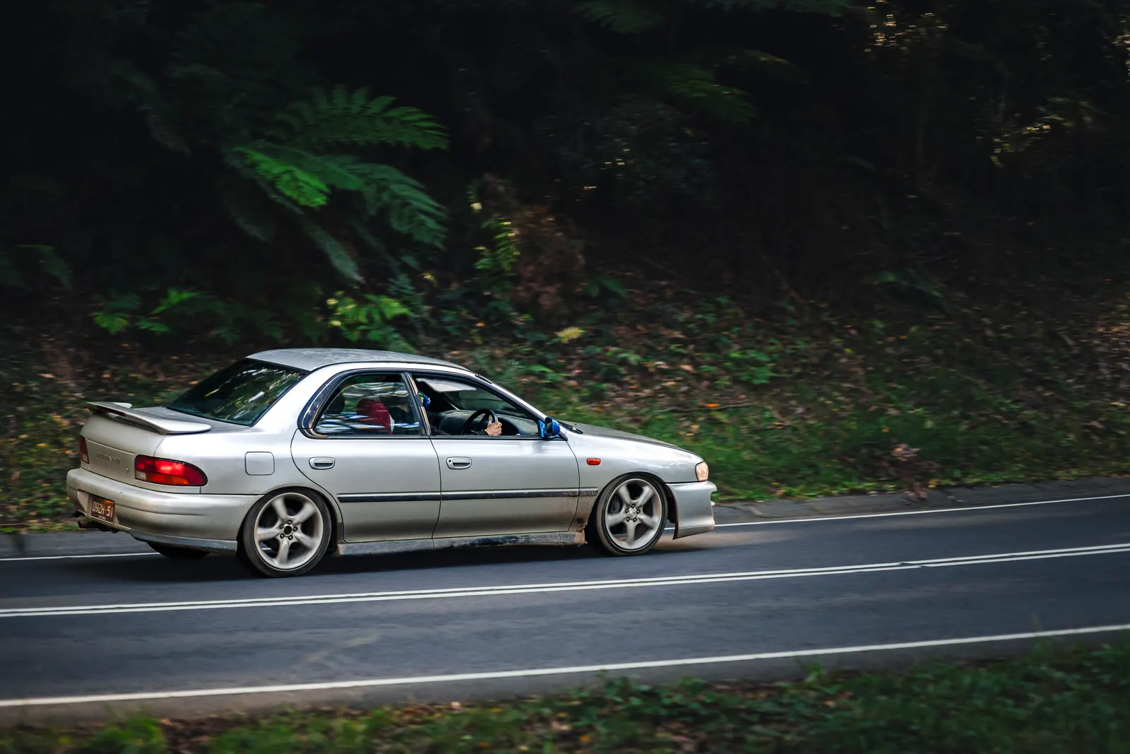 Silver sedan driving on a forested road with lush green vegetation in the background.