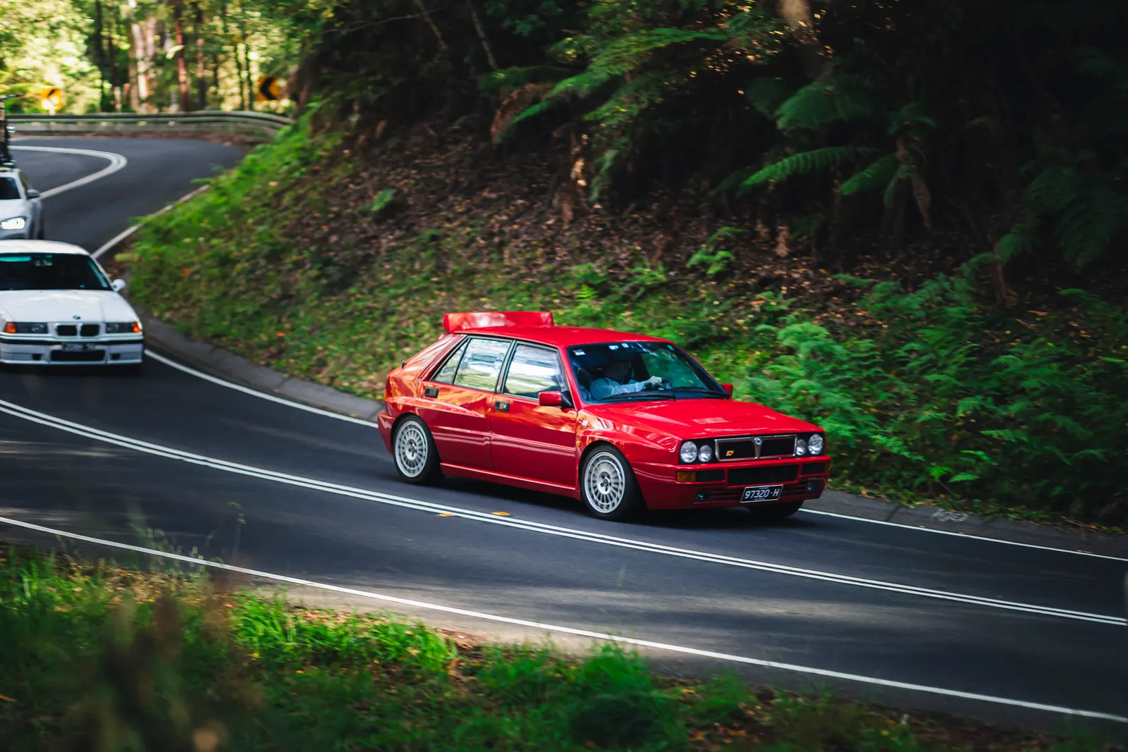 Red sedan navigating a winding forest road with dense green vegetation on the embankment.