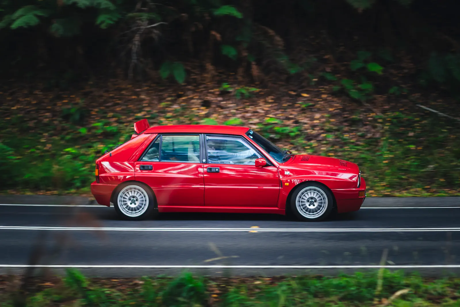 Red compact sedan driving on a two-lane road bordered by dense green forest.