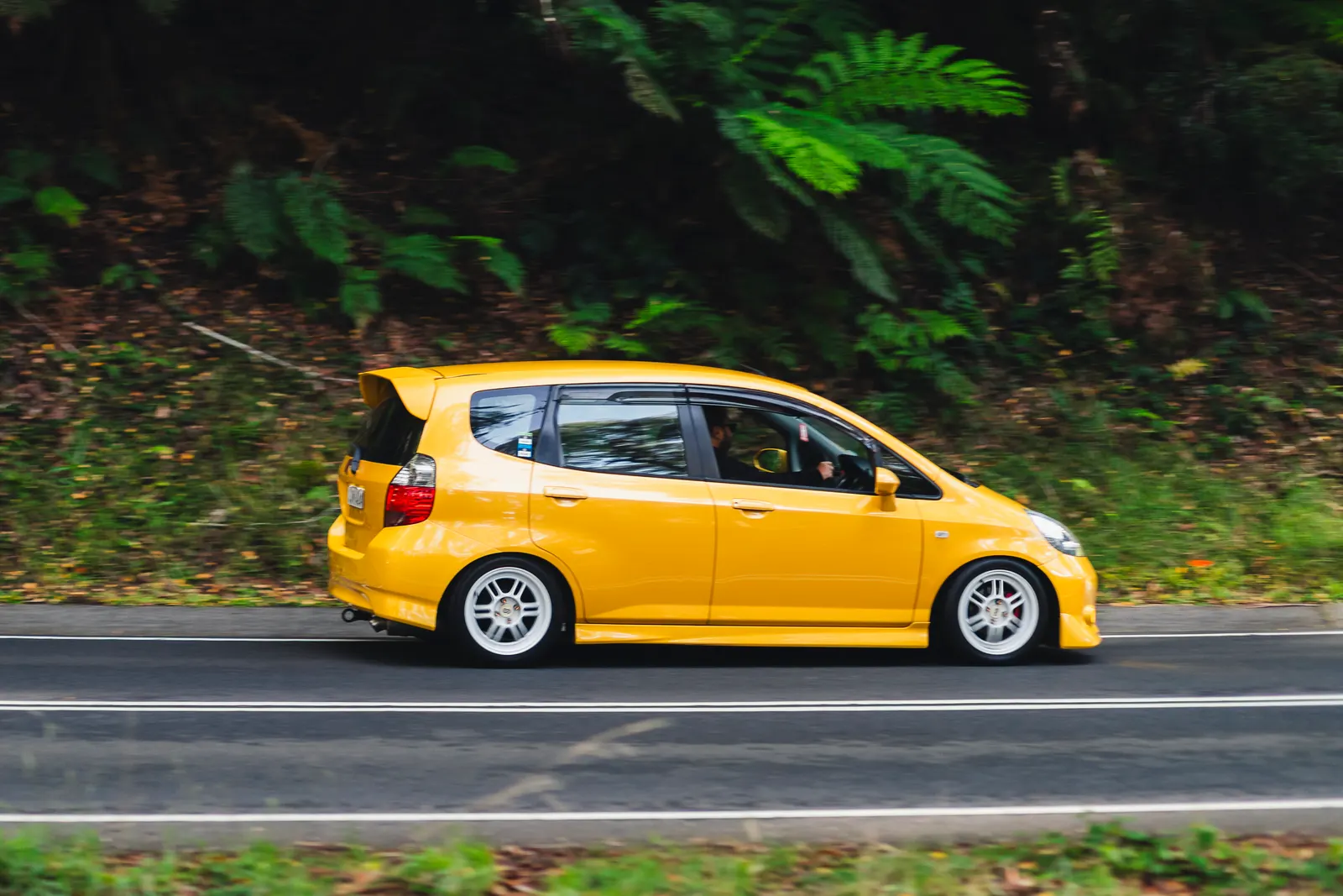 Yellow compact hatchback driving on a road bordered by dense green foliage.