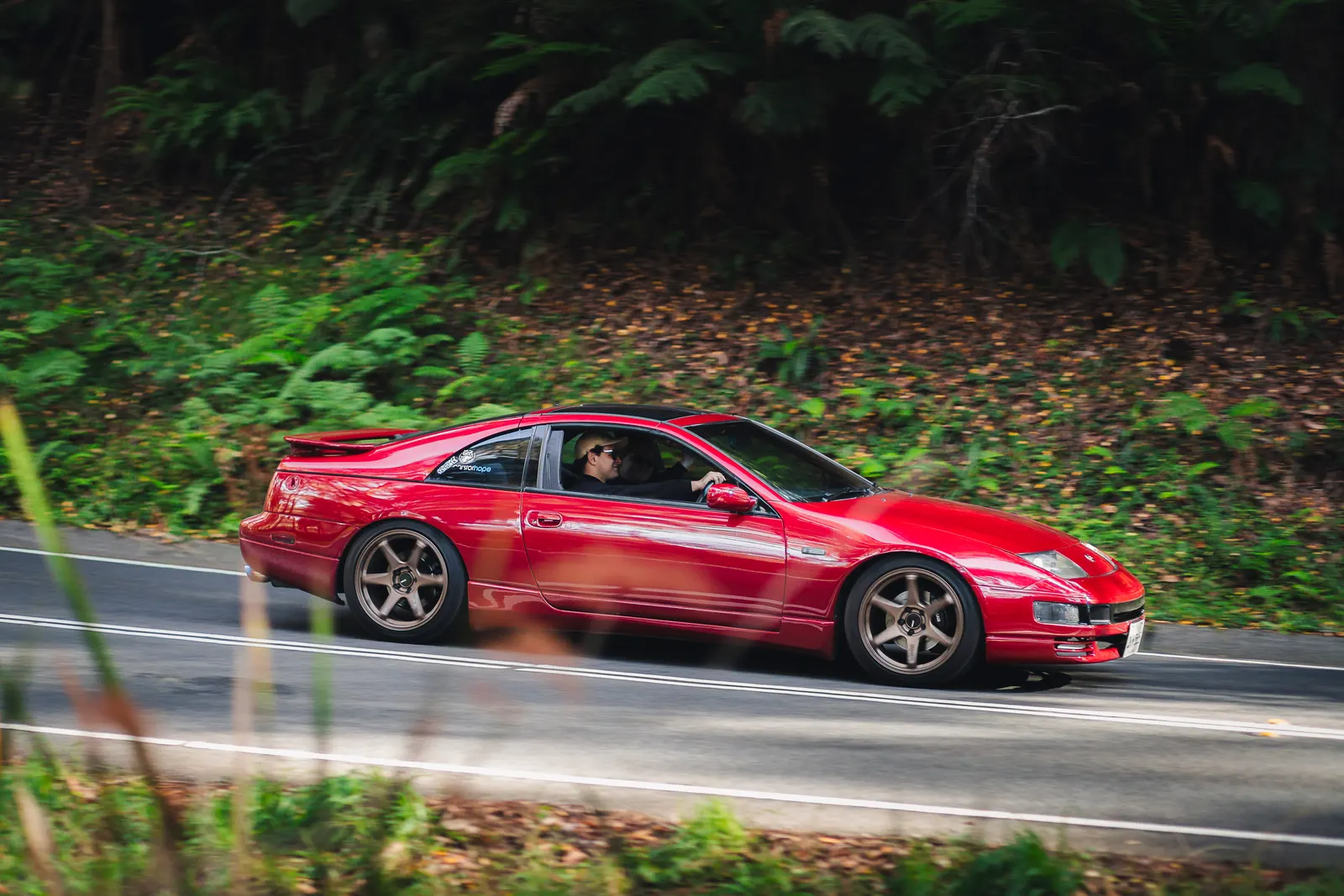 A red sports car drives along a tree-lined road with lush green foliage in the background.