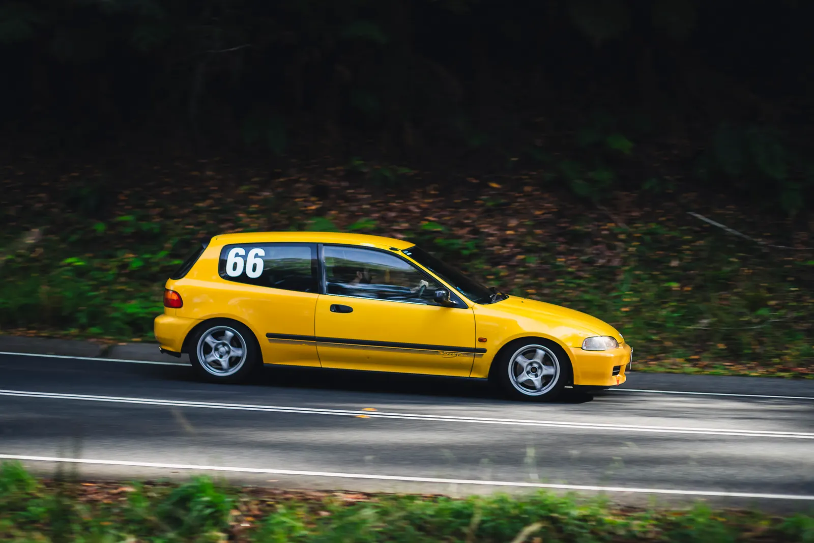 Yellow compact hatchback with racing number 66 driving on a curved road lined with dark forest.
