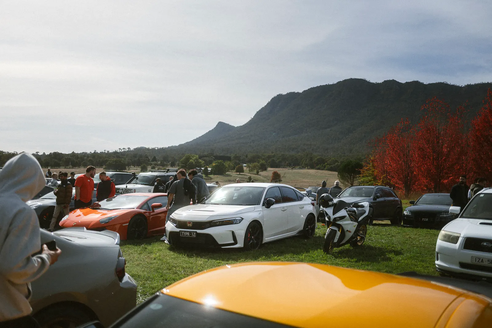 Cars and motorcycles displayed in a grass field with spectators and mountains in the background under partly cloudy sky.