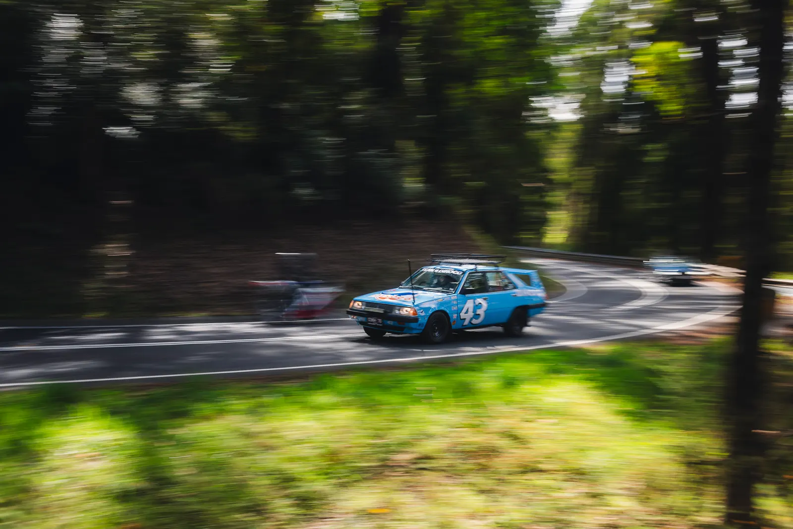 A blue and orange race car numbered 43 drives through a tree-lined road with motion blur around the foreground.